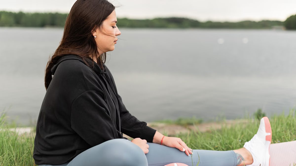 Woman meditating outdoors by a tranquil lakeside, fostering health and wellness.